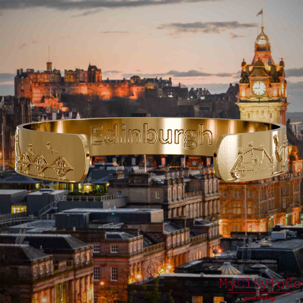 Innenseite eines vergoldeten Armreifs mit Reliefs bekannter Sehenswürdigkeiten der Stadt Edinburgh und einer Gravur des Stadtnamens. Den Hintergrund bildet eine Ansicht der Stadt Edinburgh.