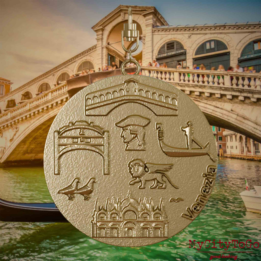 Halskette mit Drehwirbel und Vorderseite eines Schmuckanhängers aus vergoldetem Edelstahl, auf dem Reliefs bekannter Sehenswürdigkeiten der Stadt Venedig abgebildet sind. Den Hintergrund bildet eine Ansicht der Stadt Venedig.



