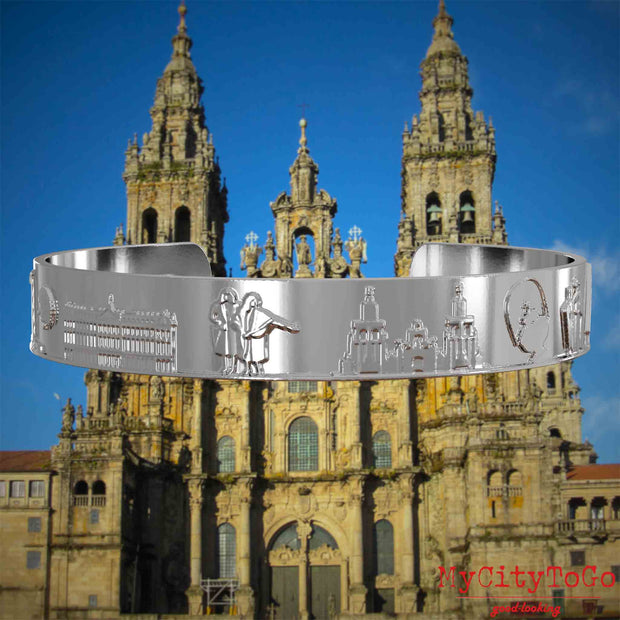 Versilberter Armreif mit Reliefs bekannter Sehenswürdigkeiten und Elemente des Jakobsweges. Den Hintergrund bildet die Kathedrale von Santiago de Compostela.