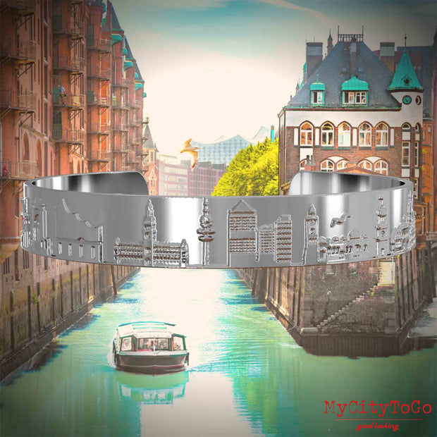 Versilberter Armreif mit Reliefs bekannter Sehenswürdigkeiten der Stadt Hamburg. Den Hintergrund bildet eine Ansicht der Stadt Hamburg.