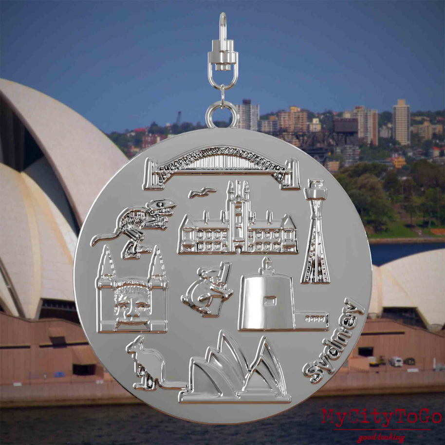 Halskette mit Drehwirbel und Vorderseite eines silbernen Schmuckanhängers, auf dem Reliefs bekannter Sehenswürdigkeiten der Stadt Sydney abgebildet sind. Den Hintergrund bildet eine Ansicht der Stadt Sydney.
