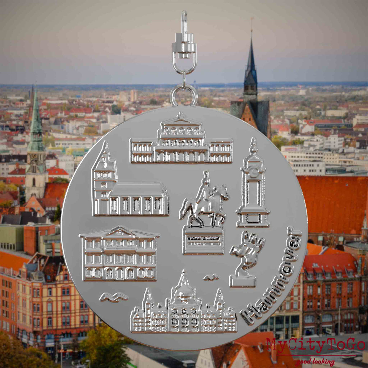 Halskette mit Drehwirbel und Rückseite eines silbernen Schmuckanhängers, auf dem Reliefs bekannter Sehenswürdigkeiten der Stadt Hannover abgebildet sind. Den Hintergrund bildet eine Ansicht der Stadt Hannover .
