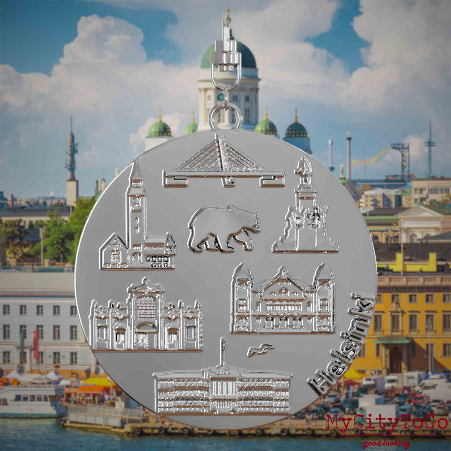 Halskette mit Drehwirbel und Rückseite eines silbernen Schmuckanhängers, auf dem Reliefs bekannter Sehenswürdigkeiten der Stadt Helsinki abgebildet sind. Den Hintergrund bildet eine Ansicht der Stadt Helsinki