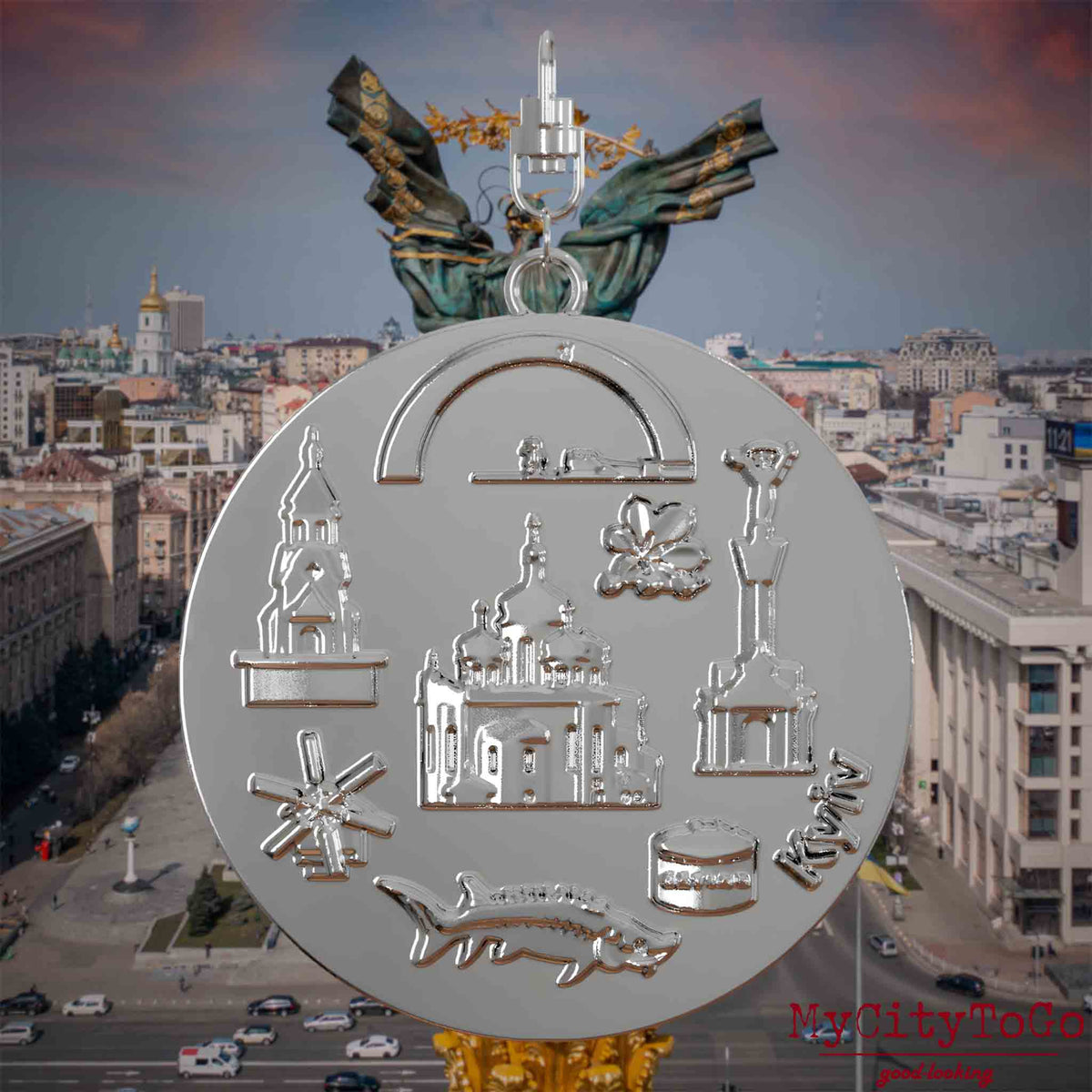Halskette mit Drehwirbel und Rückseite eines silbernen Schmuckanhängers, auf dem Reliefs bekannter Sehenswürdigkeiten der Stadt Kyjiw abgebildet sind. Den Hintergrund bildet eine Ansicht der Stadt Kyjiw.
