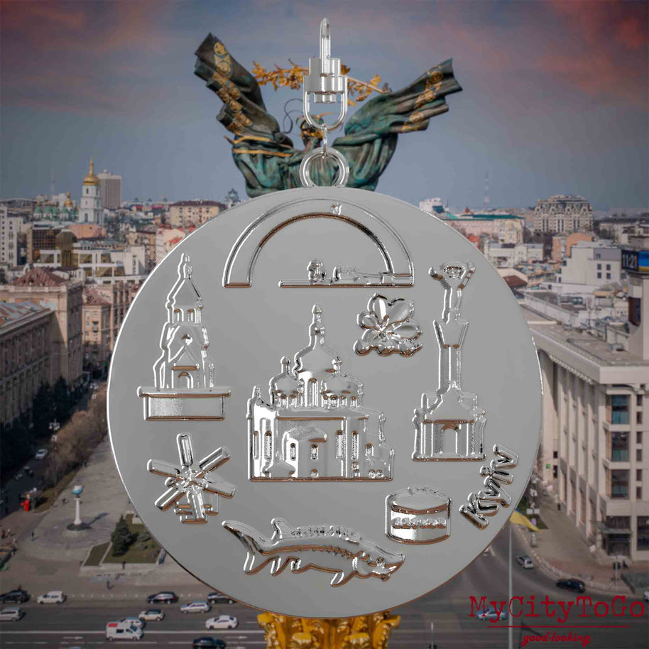 Halskette mit Drehwirbel und Rückseite eines silbernen Schmuckanhängers, auf dem Reliefs bekannter Sehenswürdigkeiten der Stadt Kyjiw abgebildet sind. Den Hintergrund bildet eine Ansicht der Stadt Kyjiw.
