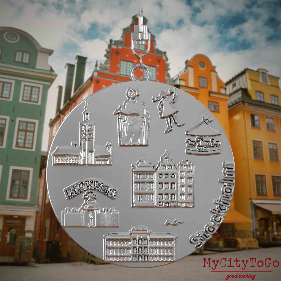 Halskette mit Drehwirbel und Rückseite eines silbernen Schmuckanhängers, auf dem Reliefs bekannter Sehenswürdigkeiten der Stadt Stockholm abgebildet sind. Den Hintergrund bildet eine Ansicht der Stadt Stockholm.
