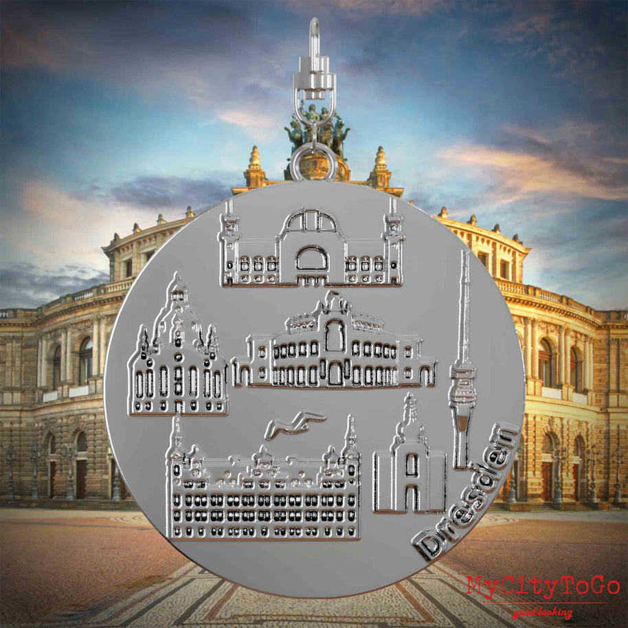 Halskette mit Drehwirbel und Rückseite eines silbernen Schmuckanhängers, auf dem Reliefs bekannter Sehenswürdigkeiten der Stadt Dresden abgebildet sind. Den Hintergrund bildet eine Ansicht der Stadt Dresden.
