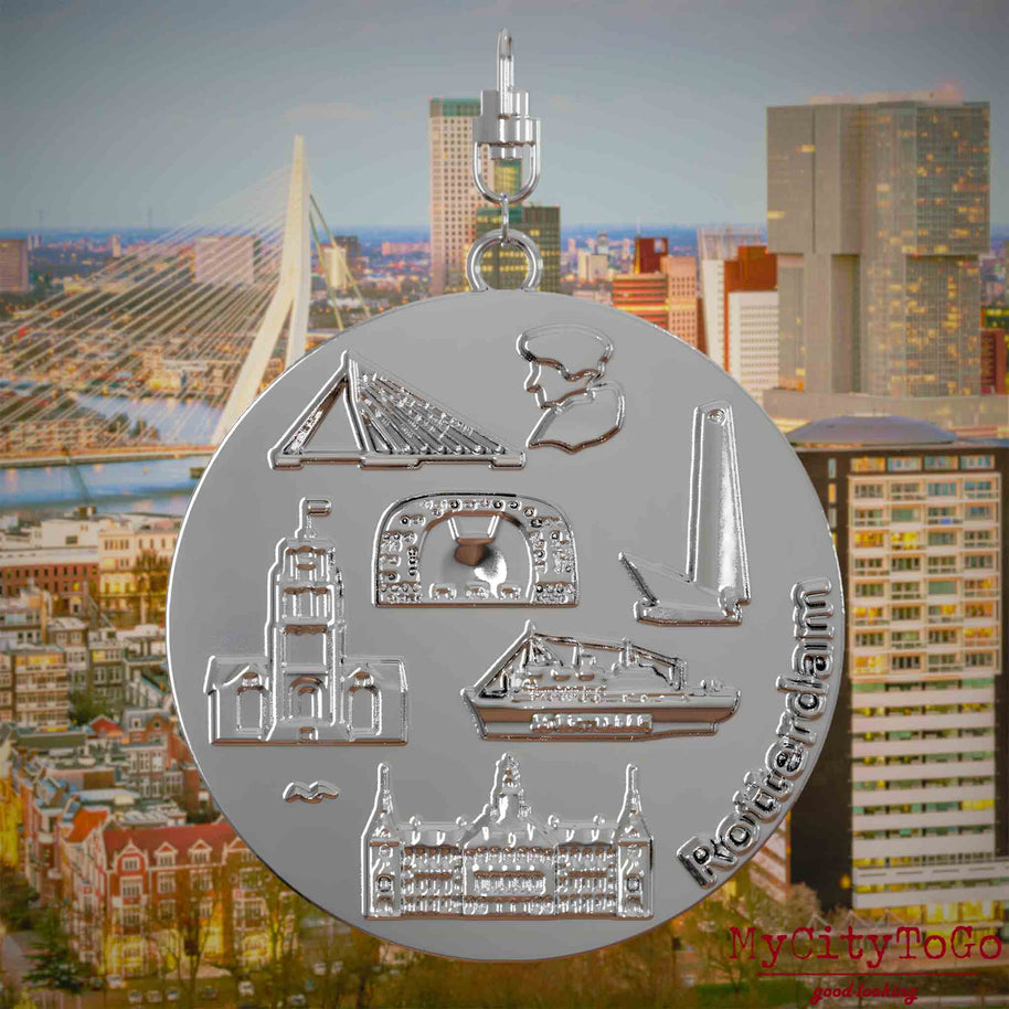 Halskette mit Drehwirbel und Rückseite eines silbernen Schmuckanhängers, auf dem Reliefs bekannter Sehenswürdigkeiten der Stadt Rotterdam abgebildet sind. Den Hintergrund bildet eine Ansicht der Stadt Rotterdam.
