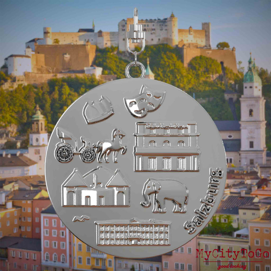 Halskette mit Drehwirbel und Rückseite eines silbernen Schmuckanhängers, auf dem Reliefs bekannter Sehenswürdigkeiten der Stadt Salzburg abgebildet sind. Den Hintergrund bildet eine Ansicht der Stadt Salzburg.

