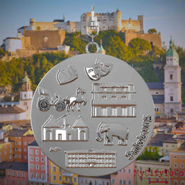 Halskette mit Drehwirbel und Rückseite eines silbernen Schmuckanhängers, auf dem Reliefs bekannter Sehenswürdigkeiten der Stadt Salzburg abgebildet sind. Den Hintergrund bildet eine Ansicht der Stadt Salzburg.

