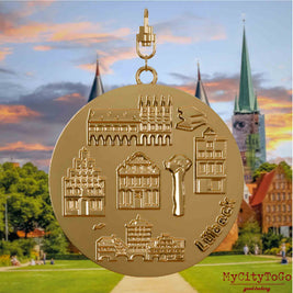 Halskette mit Drehwirbel und Vorderseite eines vergoldeten Schmuckanhängers, auf dem Reliefs bekannter Sehenswürdigkeiten der Stadt Luebeck abgebildet sind. Den Hintergrund bildet eine Ansicht der Stadt Luebeck.
