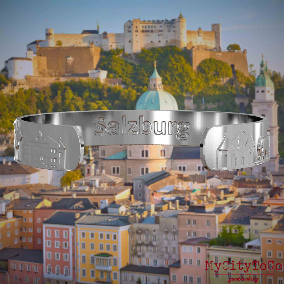 Innenseite eines silbernen Armreifs mit Reliefs bekannter Sehenswürdigkeiten der Stadt Salzburg und einer Gravur des Stadtnamens. Den Hintergrund bildet eine Ansicht der Stadt Salzburg.
