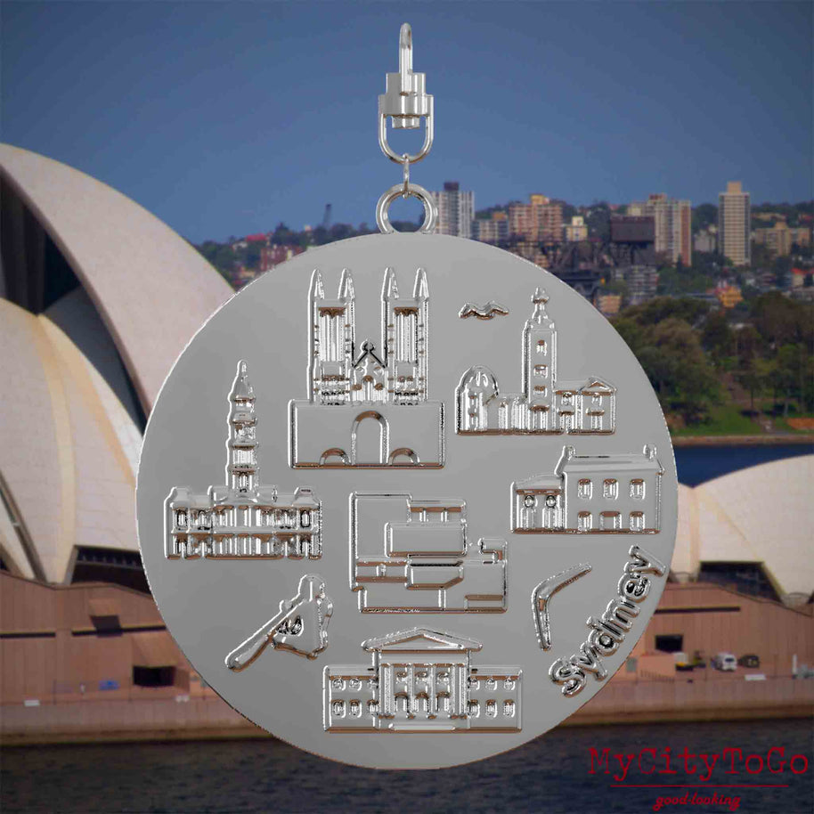 Halskette mit Drehwirbel und Rückseite eines silbernen Schmuckanhängers, auf dem Reliefs bekannter Sehenswürdigkeiten der Stadt Sydney abgebildet sind. Den Hintergrund bildet eine Ansicht der Stadt Sydney.
