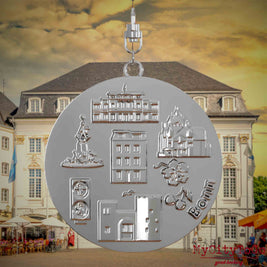 Halskette mit Drehwirbel und Rückseite eines silbernen Schmuckanhängers, auf dem Reliefs bekannter Sehenswürdigkeiten der Stadt Bonn abgebildet sind. Den Hintergrund bildet eine Ansicht der Stadt Bonn.
