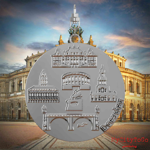Halskette mit Drehwirbel und Vorderseite eines silbernen Schmuckanhängers, auf dem Reliefs bekannter Sehenswürdigkeiten der Stadt Dresden abgebildet sind. Den Hintergrund bildet eine Ansicht der Stadt Dresden.