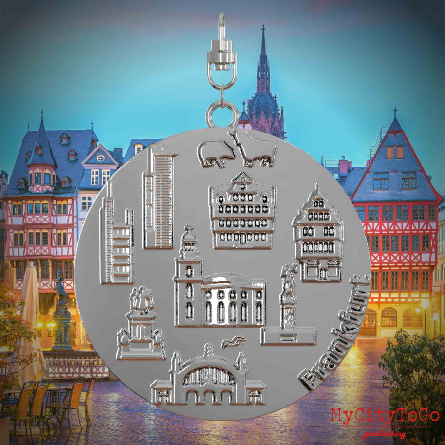 Halskette mit Drehwirbel und Vorderseite eines silbernen Schmuckanhängers, auf dem Reliefs bekannter Sehenswürdigkeiten der Stadt Frankfurt abgebildet sind. Den Hintergrund bildet eine Ansicht der Stadt Frankfurt.