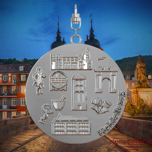 Halskette mit Drehwirbel und Vorderseite eines silbernen Schmuckanhängers, auf dem Reliefs bekannter Sehenswürdigkeiten der Stadt Heidelberg abgebildet sind. Den Hintergrund bildet eine Ansicht der Stadt Heidelberg .

