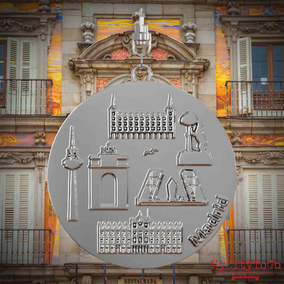 Halskette mit Drehwirbel und Vorderseite eines silbernen Schmuckanhängers, auf dem Reliefs bekannter Sehenswürdigkeiten der Stadt Madrid abgebildet sind. Den Hintergrund bildet eine Ansicht der Stadt Madrid.
