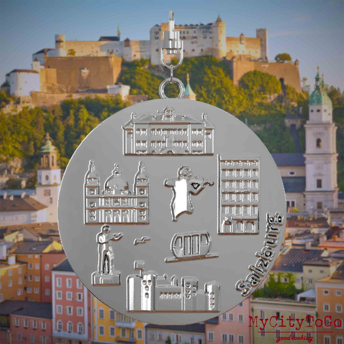 Halskette mit Drehwirbel und Vorderseite eines silbernen Schmuckanhängers, auf dem Reliefs bekannter Sehenswürdigkeiten der Stadt Salzburg abgebildet sind. Den Hintergrund bildet eine Ansicht der Stadt Salzburg.
