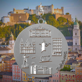 Halskette mit Drehwirbel und Vorderseite eines silbernen Schmuckanhängers, auf dem Reliefs bekannter Sehenswürdigkeiten der Stadt Salzburg abgebildet sind. Den Hintergrund bildet eine Ansicht der Stadt Salzburg.
