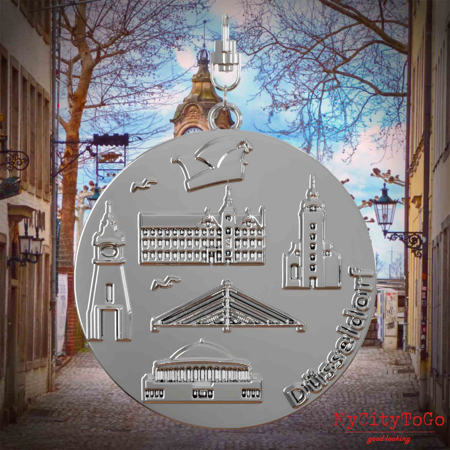 Halskette mit Drehwirbel und Rückseite eines silbernen Schmuckanhängers, auf dem Reliefs bekannter Sehenswürdigkeiten der Stadt Duesseldorf abgebildet sind. Den Hintergrund bildet eine Ansicht der Stadt Duesseldorf.