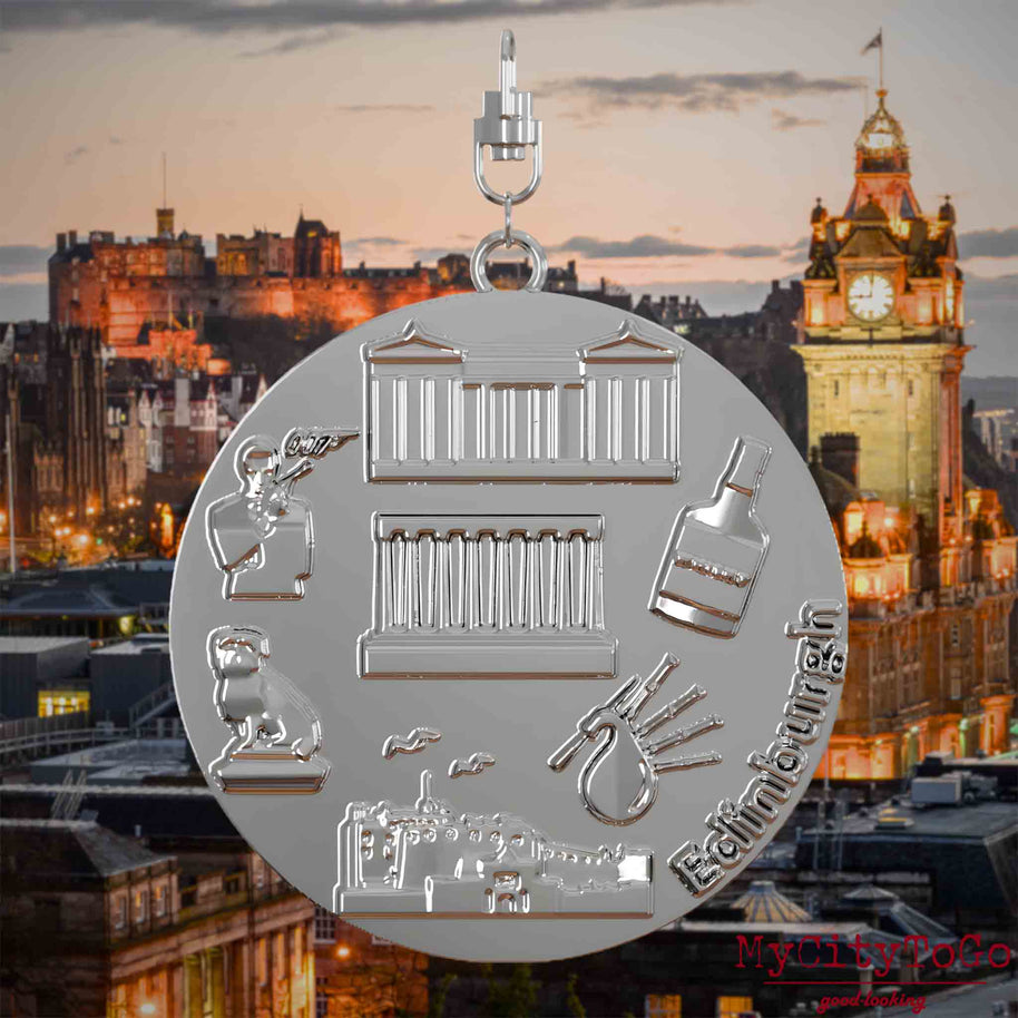 Halskette mit Drehwirbel und Vorderseite eines silbernen Schmuckanhängers, auf dem Reliefs bekannter Sehenswürdigkeiten der Stadt Edinburgh abgebildet sind. Den Hintergrund bildet eine Ansicht der Stadt Edinburgh.
