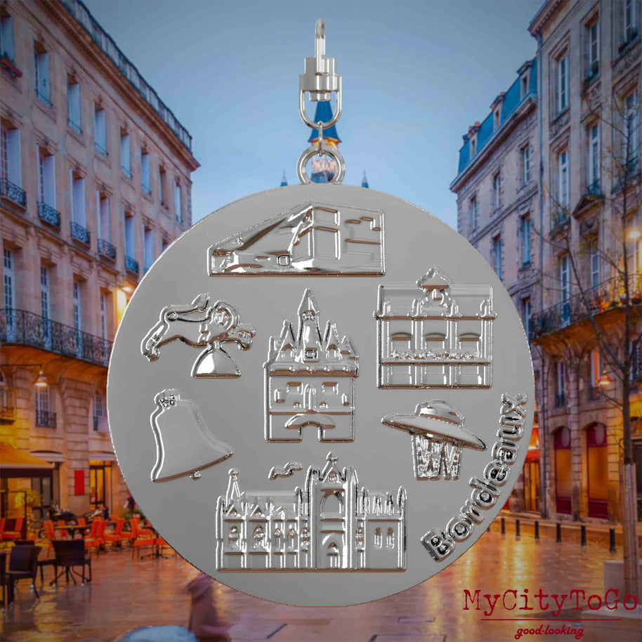 Halskette mit Drehwirbel und Rückseite eines silbernen Schmuckanhängers, auf dem Reliefs bekannter Sehenswürdigkeiten der Stadt Bordeaux abgebildet sind. Den Hintergrund bildet eine Ansicht der Stadt Bordeaux.
