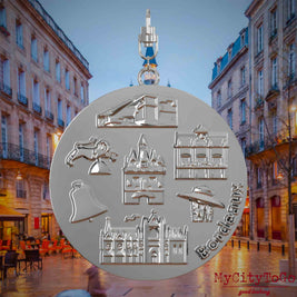 Halskette mit Drehwirbel und Rückseite eines silbernen Schmuckanhängers, auf dem Reliefs bekannter Sehenswürdigkeiten der Stadt Bordeaux abgebildet sind. Den Hintergrund bildet eine Ansicht der Stadt Bordeaux.
