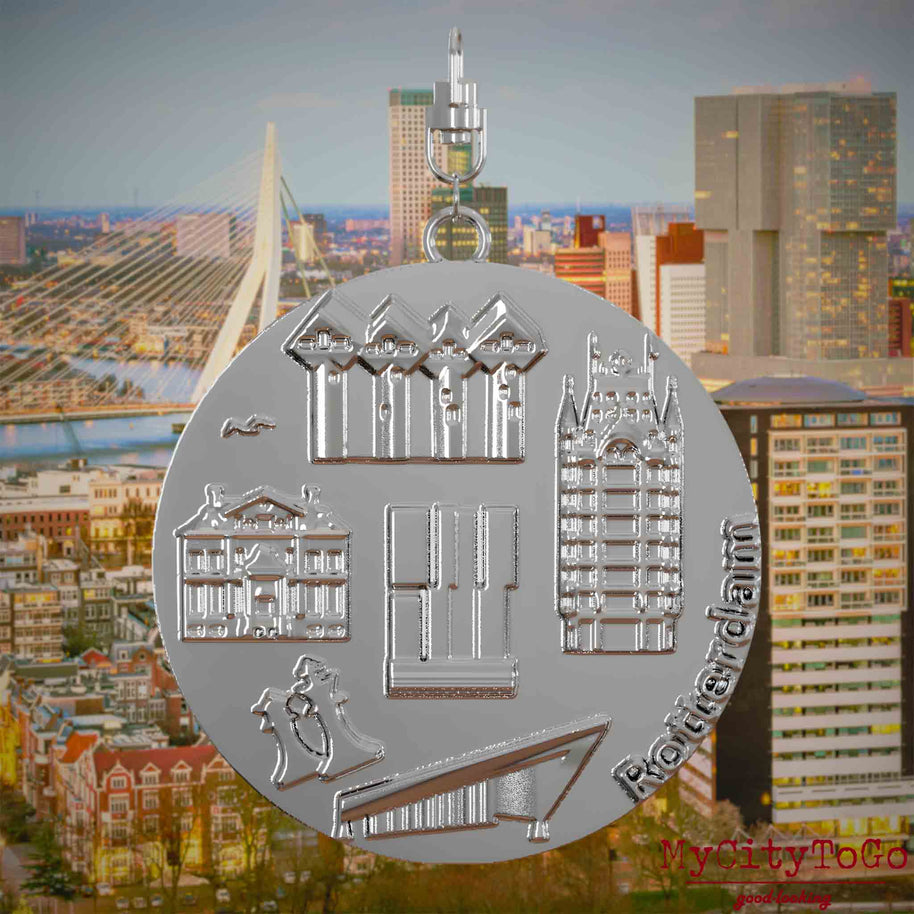 Halskette mit Drehwirbel und Vorderseite eines silbernen Schmuckanhängers, auf dem Reliefs bekannter Sehenswürdigkeiten der Stadt Rotterdam abgebildet sind. Den Hintergrund bildet eine Ansicht der Stadt Rotterdam.
