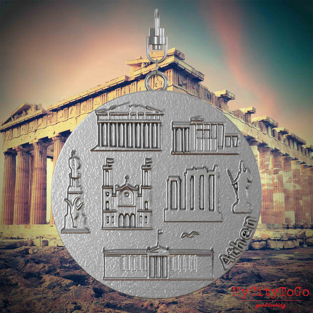 Halskette mit Drehwirbel und Vorderseite eines Schmuckanhängers aus Edelstahl, auf dem Reliefs bekannter Sehenswürdigkeiten der Stadt Athen abgebildet sind. Den Hintergrund bildet eine Ansicht der Stadt Athen.
