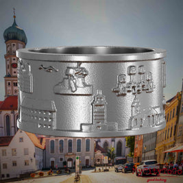 Ring aus Edelstahl mit Reliefs bekannter Sehenswürdigkeiten der Stadt Augsburg. Den Hintergrund bildet eine Ansicht der Stadt Augsburg.