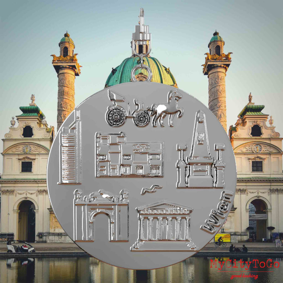 Halskette mit Drehwirbel und Vorderseite eines silbernen Schmuckanhängers, auf dem Reliefs bekannter Sehenswürdigkeiten der Stadt Wien abgebildet sind. Den Hintergrund bildet eine Ansicht der Stadt Wien.
