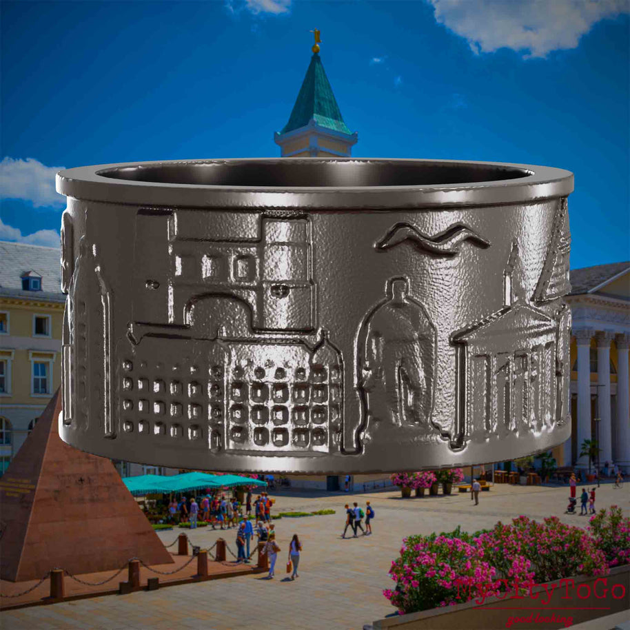 Ring aus Titan mit Reliefs bekannter Sehenswürdigkeiten der Stadt Karlsruhe. Den Hintergrund bildet eine Ansicht der Stadt Karlsruhe.