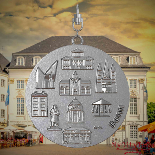 Halskette mit Drehwirbel und Vorderseite eines Schmuckanhängers aus Edelstahl, auf dem Reliefs bekannter Sehenswürdigkeiten der Stadt Bonn abgebildet sind. Den Hintergrund bildet eine Ansicht der Stadt Bonn.

