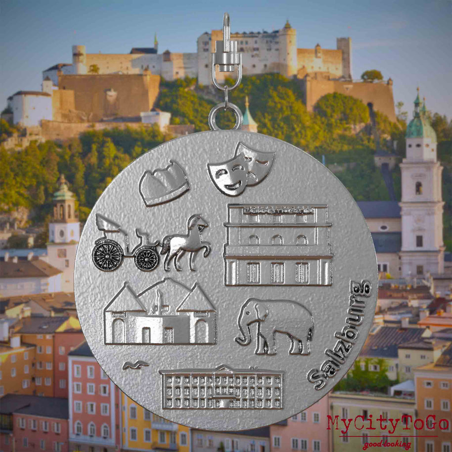 Halskette mit Drehwirbel und Rückseite eines Schmuckanhängers aus Edelstahl, auf dem Reliefs bekannter Sehenswürdigkeiten der Stadt Salzburg abgebildet sind. Den Hintergrund bildet eine Ansicht der Stadt Salzburg.

