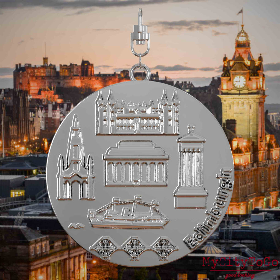 Halskette mit Drehwirbel und Rückseite eines silbernen Schmuckanhängers, auf dem Reliefs bekannter Sehenswürdigkeiten der Stadt Edinburgh abgebildet sind. Den Hintergrund bildet eine Ansicht der Stadt Edinburgh.
