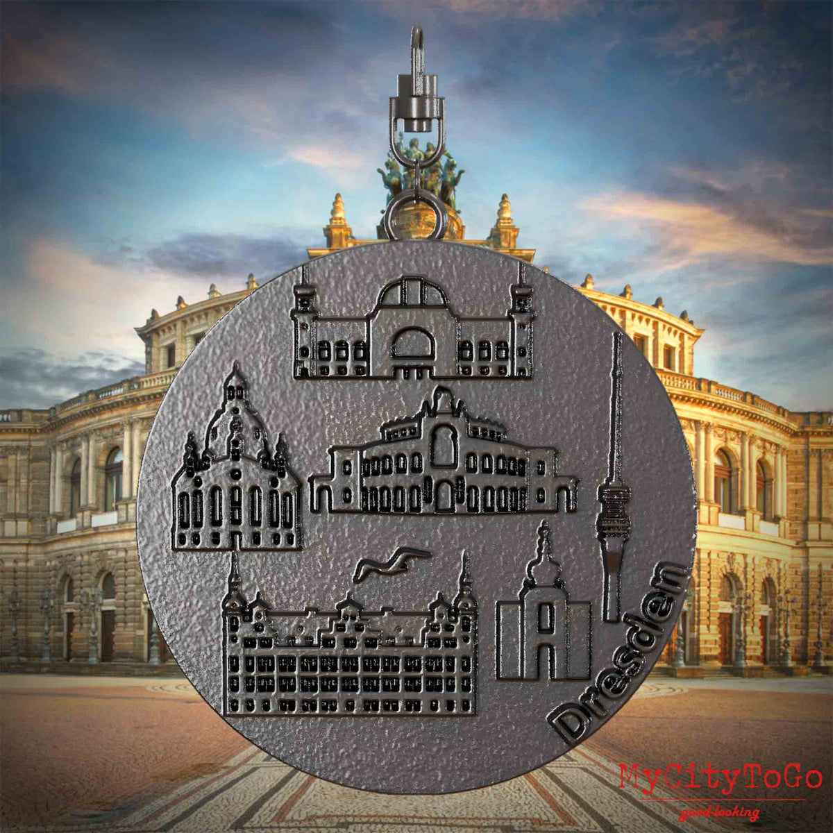 Halskette mit Drehwirbel und Rückseite eines Schmuckanhängers aus Titan, auf dem Reliefs bekannter Sehenswürdigkeiten der Stadt Dresden abgebildet sind. Den Hintergrund bildet eine Ansicht der Stadt Dresden.