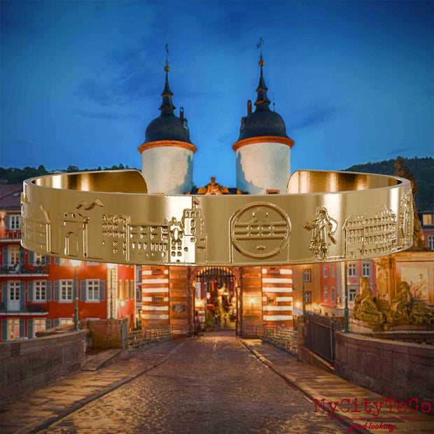 Vergoldeter Armreif mit Reliefs bekannter Sehenswürdigkeiten der Stadt Heidelberg. Den Hintergrund bildet eine Ansicht der Stadt Heidelberg.