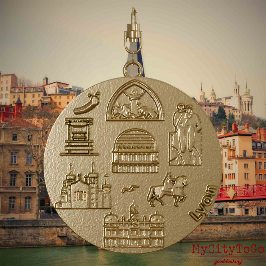 Halskette mit Drehwirbel und Rückseite eines Schmuckanhängers aus vergoldetem Edelstahl, auf dem Reliefs bekannter Sehenswürdigkeiten der Stadt Lyon abgebildet sind. Den Hintergrund bildet eine Ansicht der Stadt Lyon.
