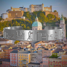 Armreif aus Edelstahl mit Reliefs bekannter Sehenswürdigkeiten der Stadt Salzburg. Den Hintergrund bildet eine Ansicht der Stadt Salzburg.