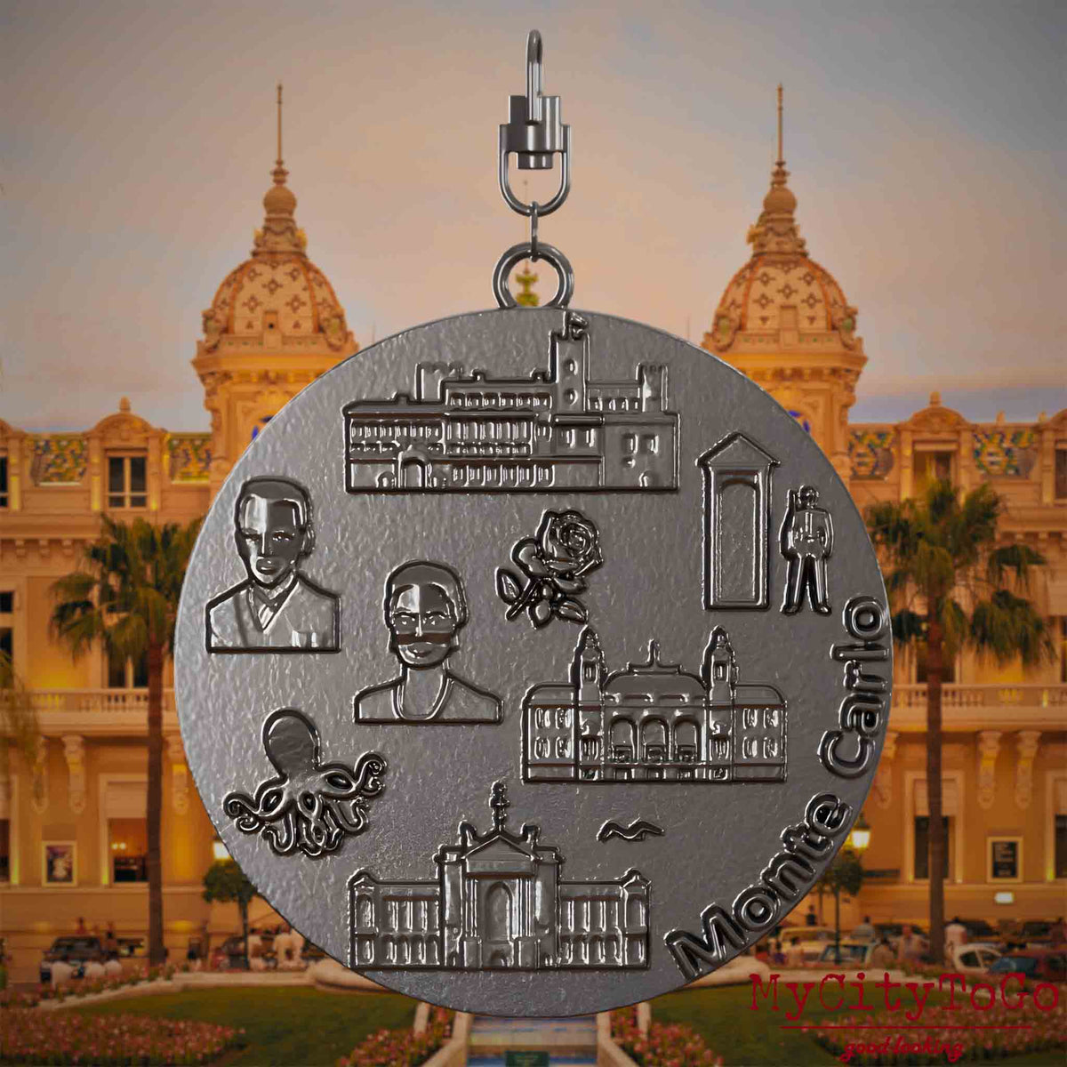 Halskette mit Drehwirbel und Rückseite eines Schmuckanhängers aus Titan, auf dem Reliefs bekannter Sehenswürdigkeiten der Stadt Monte Carlo abgebildet sind. Den Hintergrund bildet eine Ansicht der Stadt Monte Carlo.
