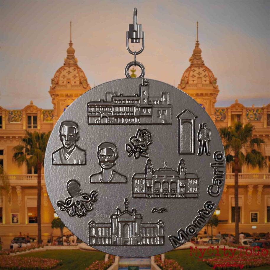 Halskette mit Drehwirbel und Rückseite eines Schmuckanhängers aus Titan, auf dem Reliefs bekannter Sehenswürdigkeiten der Stadt Monte Carlo abgebildet sind. Den Hintergrund bildet eine Ansicht der Stadt Monte Carlo.
