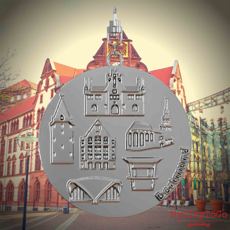 Halskette mit Drehwirbel und Rückseite eines silbernen Schmuckanhängers, auf dem Reliefs bekannter Sehenswürdigkeiten der Stadt Dortmund abgebildet sind. Den Hintergrund bildet eine Ansicht der Stadt Dortmund.