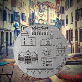 Halskette mit Drehwirbel und Rückseite eines Schmuckanhängers aus Edelstahl, auf dem Reliefs bekannter Sehenswürdigkeiten der Stadt Zuerich abgebildet sind. Den Hintergrund bildet eine Ansicht der Stadt Zuerich.
