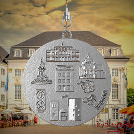 Halskette mit Drehwirbel und Rückseite eines Schmuckanhängers aus Edelstahl, auf dem Reliefs bekannter Sehenswürdigkeiten der Stadt Bonn abgebildet sind. Den Hintergrund bildet eine Ansicht der Stadt Bonn.

