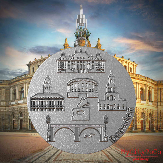 Halskette mit Drehwirbel und Vorderseite eines Schmuckanhängers aus Edelstahl, auf dem Reliefs bekannter Sehenswürdigkeiten der Stadt Dresdenabgebildet sind. Den Hintergrund bildet eine Ansicht der Stadt Dresden.