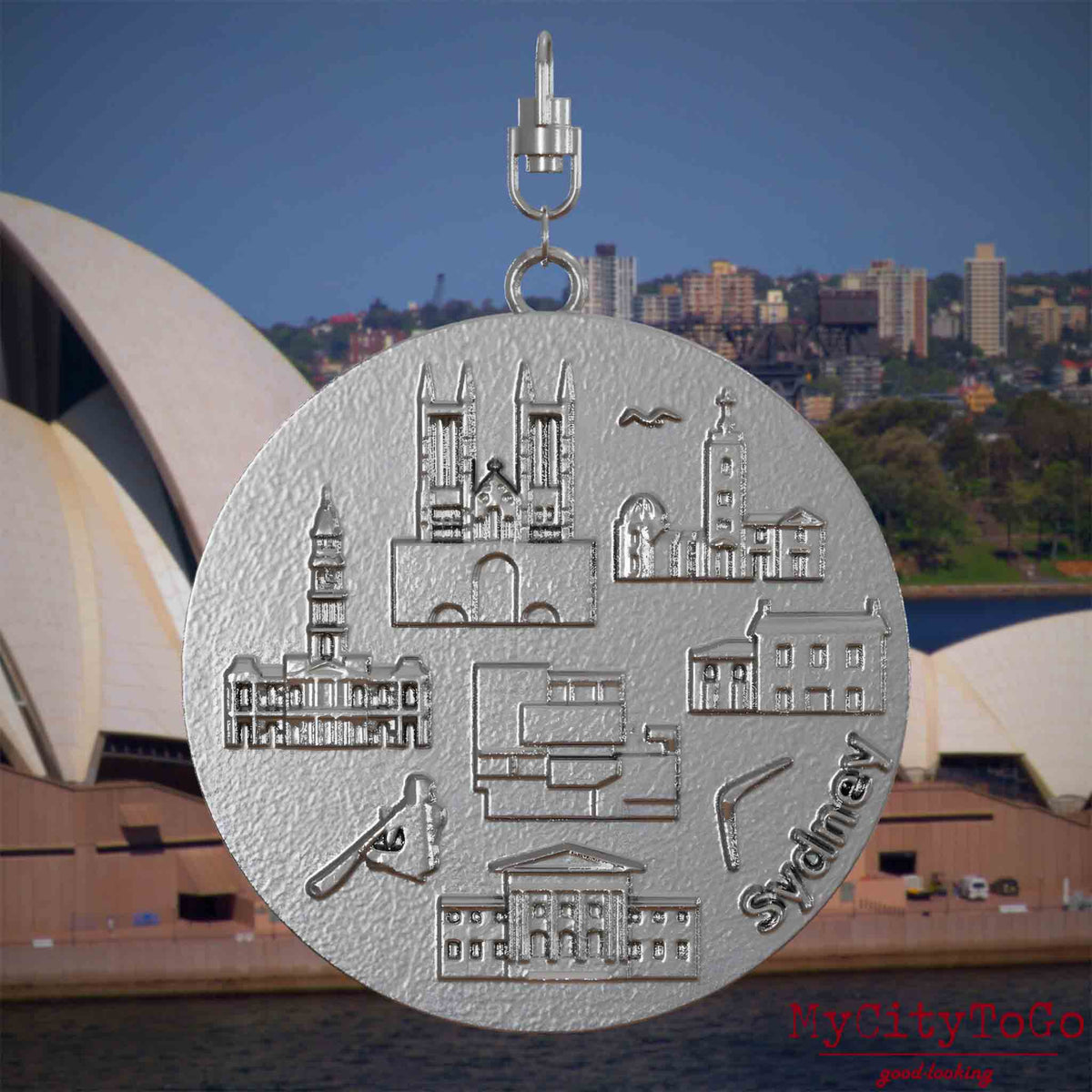 Halskette mit Drehwirbel und Rückseite eines Schmuckanhängers aus Edelstahl, auf dem Reliefs bekannter Sehenswürdigkeiten der Stadt Sydney abgebildet sind. Den Hintergrund bildet eine Ansicht der Stadt Sydney.

