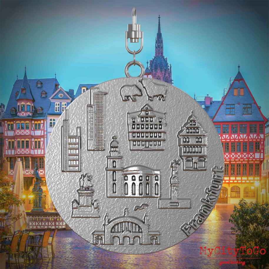 Halskette mit Drehwirbel und Vorderseite eines Schmuckanhängers aus Edelstahl, auf dem Reliefs bekannter Sehenswürdigkeiten der Stadt Frankfurt abgebildet sind. Den Hintergrund bildet eine Ansicht der Stadt Frankfurt.
