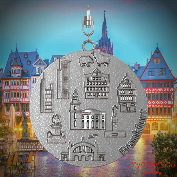 Halskette mit Drehwirbel und Vorderseite eines Schmuckanhängers aus Edelstahl, auf dem Reliefs bekannter Sehenswürdigkeiten der Stadt Frankfurt abgebildet sind. Den Hintergrund bildet eine Ansicht der Stadt Frankfurt.
