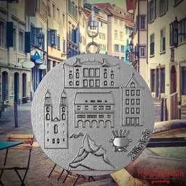Halskette mit Drehwirbel und Vorderseite eines Schmuckanhängers aus Edelstahl, auf dem Reliefs bekannter Sehenswürdigkeiten der Stadt Zuerich abgebildet sind. Den Hintergrund bildet eine Ansicht der Stadt Zuerich.
